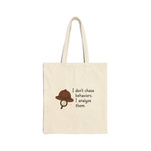 Puede incluir: Bolso tote de color crema con un sombrero de detective marrón y una lupa. El texto dice: "No persigo comportamientos. Los analizo." El bolso tiene un diseño sencillo con asas largas, adecuado para llevar libros o compras.