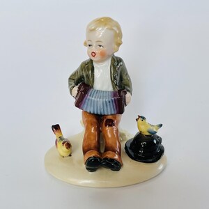 Könnte beinhalten: Eine Porzellanfigur eines Kindes mit blonden Haaren, das eine grüne Jacke und eine braune Hose trägt und ein blaues Akkordeon spielt. Das Kind sitzt auf einem Sockel mit einem kleinen gelben Vogel links und einem schwarzen Hut rechts.