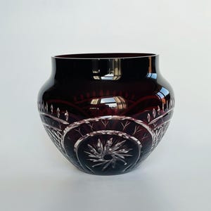 Può includere: Un vaso in vetro color bordeaux profondo con intricati motivi in vetro tagliato. Il design include un motivo a stella prominente al centro, circondato da linee arcuate e dettagli a tratteggio incrociato. Il vetro ha un bordo e una base lisci.