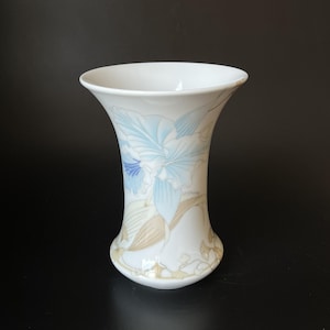 May include: A white porcelain vase with a flared rim, featuring a delicate illustration of a blue orchid flower and tan foliage. The name LEONARD is printed in small black letters on the side of the floral design, set against a smooth surface.