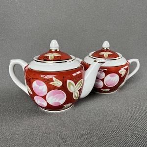 Pode incluir: Um bule e um açucareiro a condizer em cerâmica, ambos com uma cor base vermelho profundo e detalhes em branco. As peças são decoradas com motivos florais circulares cor-de-rosa pintados à mão e folhas verdes, com bordos dourados nas tampas.