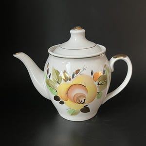 Könnte beinhalten: Eine weiße Keramik-Teekanne mit einem handgemalten gelben Rosenmotiv, grünen Blättern und goldenen Akzenten. Die Teekanne verfügt über einen passenden Deckel mit goldenem Knauf, einen geschwungenen Ausguss und einen Henkel mit Goldverzierungen.