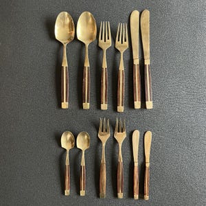 Op de afbeelding: Een set van twaalf bestekdelen met goudkleurige metalen koppen en donkerbruine houten handvatten. De collectie bevat twee grote lepels, twee vorken, twee messen, twee kleine lepels, twee kleine vorken en twee kleine messen, gerangschikt in rijen.