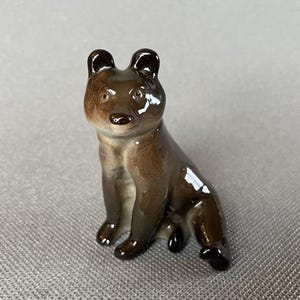 May include: A small, brown ceramic bear figurine in a seated position. The bear has dark brown ears, nose, and paws, with a glossy finish. The figurine is a decorative item, likely for display.