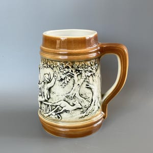 Peut inclure: Une chope à bière en céramique avec un bord et une base émaillés marron, présentant un relief couleur crème représentant une personne coiffée d'un chapeau assise dans une forêt avec un petit animal. La chope a une anse robuste.