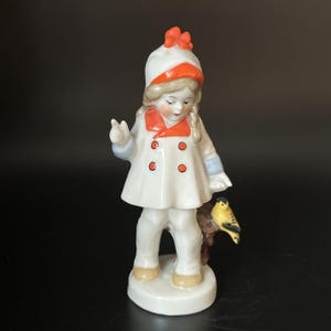 Może przedstawiać: Porcelanowa figurka dziecka ubranego w biały płaszcz z pomarańczowymi guzikami, pomarańczowym kołnierzem i pasującą białą czapką z pomarańczowym rondem. Dziecko ma jasnobrązowe włosy zaplecione w warkocze i stoi na białej podstawie obok małego żółtego ptaszka.
