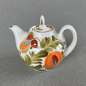 Op de afbeelding: Een witte porseleinen theepot met een handgeschilderd bloemmotief in olijfgroen, feloranje en turquoise. De theepot heeft een bijpassend deksel met knop, een gebogen handvat en een tuit, allemaal afgewerkt met een gouden randje.