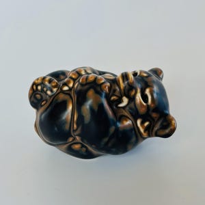 Può includere: Una piccola statuetta in ceramica di un animale raggomitolato con una finitura lucida. La superficie presenta un motivo marmorizzato nei toni del nero profondo e del marrone ambra caldo, creando una forma liscia e arrotondata con contorni.