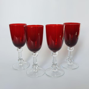 Può includere: Un set di quattro calici da vino abbinati con coppe rosso intenso e steli in vetro trasparente. Ogni calice presenta una forma arrotondata, un nodo sferico distintivo sullo stelo e una base circolare ampia e piatta per la stabilità.
