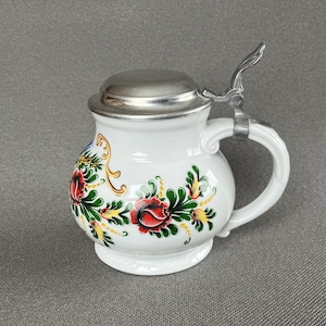 Op de afbeelding: Een witte keramische bierpul met een ronde buik en een scharnierend metalen deksel. De buitenkant is voorzien van handgeschilderde bloemmotieven in rood, groen en geel, met goudkleurige krulaccenten. Het heeft een gebogen handvat en een duimhendel.