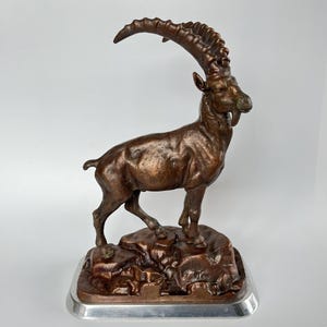 Könnte beinhalten: Eine bronzefarbene Skulptur eines Steinbocks, der auf einem felsigen Untergrund steht. Das Tier hat lange, gebogene, geriffelte Hörner, einen kurzen Bart und einen muskulösen Körperbau. Die Figur steht auf einem rechteckigen, silberfarbenen Sockel.