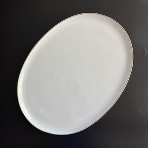 Puede incluir: Una bandeja de servir ovalada, blanca y lisa, que presenta un borde fino de color dorado metálico a lo largo del contorno. La superficie de la bandeja es brillante y reflectante, ofreciendo una base limpia y neutra para presentar alimentos.