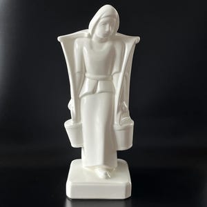 May include: A white ceramic figurine of a woman carrying two buckets. The figure is wearing a long dress and a head covering. The sculpture has a rectangular base and is set against a black background.
