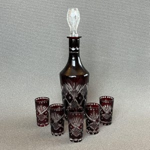 Puede incluir: Una jarra de cristal rojo intenso con un tapón de cristal transparente y cinco vasos altos a juego. Cada pieza presenta intrincados patrones de cristal tallado, incluyendo motivos de rejilla y estrellas, contrastando el rojo oscuro y el cristal.