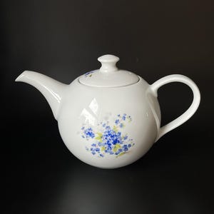Può includere: Una teiera in ceramica bianca con corpo arrotondato, manico curvo e beccuccio. La teiera presenta un delicato motivo floreale di piccoli fiori blu con foglie verdi sulla parte anteriore e un dettaglio floreale coordinato sul pomello del coperchio.