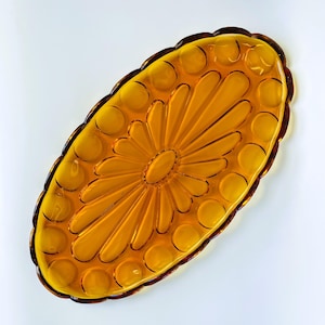 Op de afbeelding: Een ovaal, amberkleurig glazen dienblad met een geschulpte rand en een decoratief patroon van verhoogde cirkelvormige stippen langs de omtrek, rondom een centraal zonnestraalontwerp met langwerpige bloemblaadjes vanuit een glad, ovaal middelpunt.