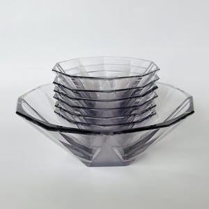 Puede incluir: Un conjunto de cuencos de cristal gris translúcido apilados con un diseño geométrico facetado de múltiples lados. Los cuencos tienen un borde ancho e inclinado y una base cónica, creando una apariencia prismática en capas con bordes definidos.