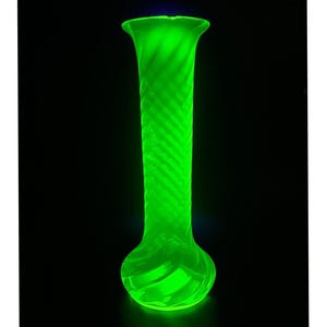 Puede incluir: Un florero de vidrio alto y esbelto con un tono verde neón brillante y resplandeciente. La superficie del florero muestra un patrón de espiral diagonal distintivo y repetitivo que envuelve todo el cuerpo, desde el borde hasta la base.