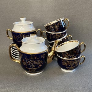 Op de afbeelding: Een theeservies bestaande uit een theepot, een suikerpot met deksel en diverse theekopjes met schoteltjes. Elk onderdeel is gemaakt van wit porselein met een diep marineblauwe basis, versierd met gouden bloemmotieven en gouden randen en oren.