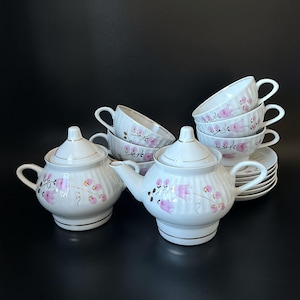 Pode incluir: Um conjunto de chá em porcelana branca com textura canelada e um delicado padrão floral rosa com acabamento dourado. O conjunto inclui um bule com tampa, um açucareiro com tampa e várias chávenas de chá com pires a condizer em grupo.