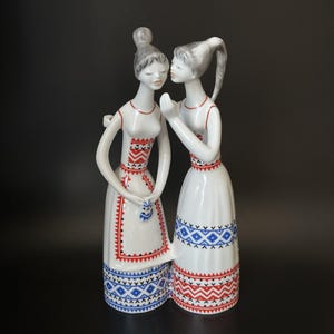 Può includere: Una statuetta in porcellana bianca raffigurante due figure in piedi l'una accanto all'altra, una delle quali sussurra all'altra. Entrambe indossano abiti lunghi decorati con intricati motivi geometrici rossi e blu. Hanno capelli grigi raccolti in chignon.