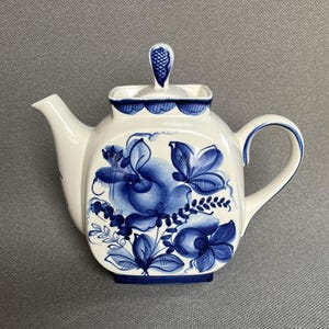 Può includere: Una teiera in ceramica di forma quadrata con base bianca e motivi floreali blu. La teiera presenta un coperchio abbinato con un pomello decorativo, un manico curvo e un beccuccio. Il design blu include petali e foglie dettagliate.