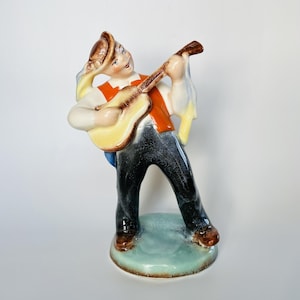 Pode incluir: Uma estatueta de cerâmica de um músico com chapéu castanho, colete laranja e calças azul-escuras, a tocar uma guitarra acústica amarelo-clara. A figura assenta numa base redonda azul-clara com acabamento brilhante e rebordo castanho.