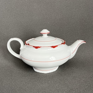 Op de afbeelding: Een witte porseleinen theepot met een ronde vorm, een gebogen handvat en een bijpassend deksel met een kleine knop. De theepot is versierd met dunne, horizontale rode lijnen en een geschulpte rode rand langs de bovenkant van de pot.