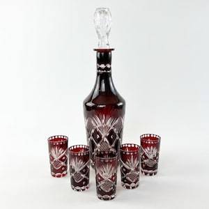 May include: A deep red cut-to-clear crystal decanter with a matching clear glass stopper and five coordinating tall drinking glasses. Each piece features intricate diamond-shaped cross-hatching and starburst patterns cut into the dark red glass.
