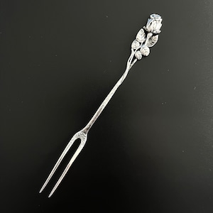 Könnte beinhalten: Eine silberfarbene Cocktailgabel aus Metall mit einem langen, schlanken Griff, der in einer detaillierten, dreidimensionalen Rosenknospe endet, ergänzt durch strukturierte Blätter und ein stängelartiges Design entlang des gesamten Bestecks.