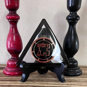 May include: A black triangular display stand holding a circular emblem with a red and gold design. The emblem is set against a black background. Two wooden candle holders, one red and one black, flank the display.