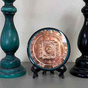 May include: A copper-colored mandala with intricate designs and a turquoise border is displayed on a black stand. The mandala is surrounded by a ring of text in a foreign language.