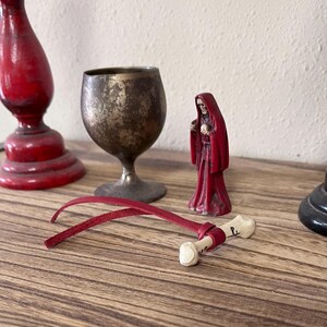 May include: A small, off-white bone-shaped object with a black symbol, wrapped in a red leather cord. Nearby are a red candle holder, a tarnished brass goblet, and a small red-robed figurine with a skeletal face standing on a wooden surface.