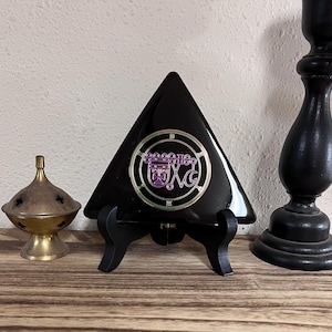 May include: A black triangular decorative piece with a purple and gold emblem, displayed on a black stand. Beside it is a brass incense burner and a black candlestick holder. The items are on a wooden surface.