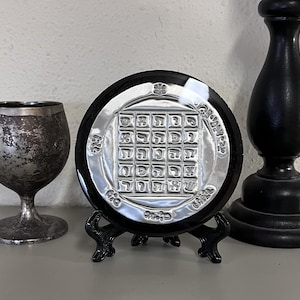 May include: A silver-coloured, round decorative plate with an intricate design, displayed on a black stand. The plate has a black border and is flanked by a silver goblet and a black candlestick holder.