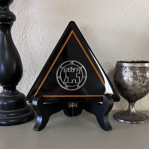 May include: A black triangular glass plate with a gold border and a silver sigil in the center. The plate is on a black stand.