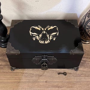 May include: A rectangular black wooden box featuring a light-colored engraved skull design on the lid. The box includes dark metal corner protectors, a decorative front latch with a small padlock, and claw-shaped feet. A small metal key lies nearby.