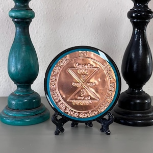 Puede incluir: Un objeto redondo de color cobre con diseños y texto intrincados, enmarcado en negro con un borde turquesa, exhibido en un pequeño soporte negro. Dos candelabros decorativos turquesa y negros flanquean el objeto, creando una composición simétrica.