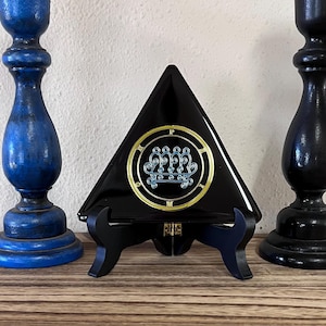 May include: A black triangular decorative piece with a gold and blue emblem, displayed on a black stand. The emblem features intricate designs within a circular frame. Two blue candle holders flank the piece, adding a touch of contrast.