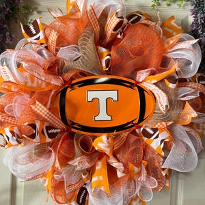 May include: A decorative wreath featuring orange, white, and brown mesh, ribbons, and football accents. The wreath has a central orange football-shaped emblem with a white "T" logo, perfect for sports fans.