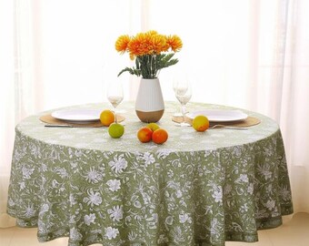 Può includere: Una tovaglia rotonda verde con un motivo floreale bianco copre un tavolo di legno scuro. La tavola è apparecchiata con piatti, bicchieri e un vaso di fiori arancioni. Diversi frutti sono posti sul tavolo.
