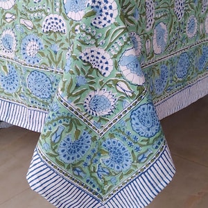 May include: A green floral tablecloth with a repeating pattern of blue and white flowers and green leaves. The border features blue and white stripes. The tablecloth is draped over a wooden table, showcasing the design and texture.
