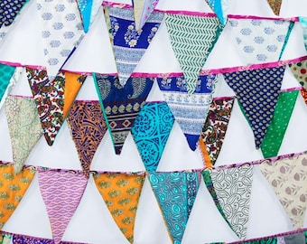 May include: A colorful bunting garland with triangular flags. The flags feature various patterns and colors, including white, blue, orange, green, and pink. The garland is trimmed with pink ribbon and is suitable for festive decorations.