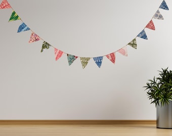 May include: A colorful fabric banner with triangular flags in various patterns and colors, including red, blue, green, and pink, hangs against a white wall. The banner is suspended above a wooden floor, with a potted plant in the corner.