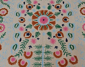 Puede incluir: Textil beige con un patrón floral simétrico. El diseño presenta grandes flores estilizadas en tonos marrón, rosa y azul, con hojas y acentos verdes. La estética general recuerda al bordado tradicional.