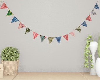 May include: A colorful fabric banner with triangular flags in various patterns and colors, strung along a thin brown cord. The flags include red, green, blue, and pink designs. The banner is hanging against a neutral wall.