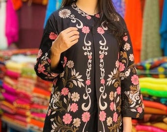 May include: Black embroidered coat with pink and white floral designs. The coat features a round collar and three-quarter sleeves. The floral embroidery is detailed and covers the front and sleeves of the coat. The background shows colorful textiles.