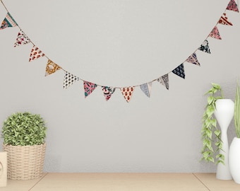 May include: A decorative garland of small, colorful fabric pennants strung on a thin cord. The pennants feature various patterns, including floral and geometric designs, in shades of red, pink, blue, and yellow. The garland is hanging against a neutral wall.