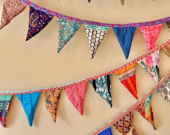 May include: Colorful fabric bunting with triangular flags in various patterns and colors, including pink, blue, orange, and green. The bunting is strung together with a decorative trim, creating a festive and decorative display.