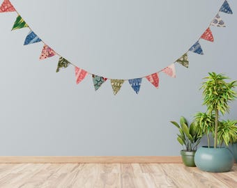 May include: A colorful fabric banner with triangular flags hangs against a light blue wall. The flags feature various patterns in red, blue, green, and pink. The banner is strung on a natural rope, adding a rustic touch to the festive decoration.